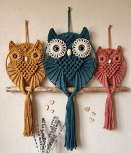 Three macramé owls in teal, mustard, and dusty rose mounted on a driftwood branch, showing the finished owl family trio macramé pattern.